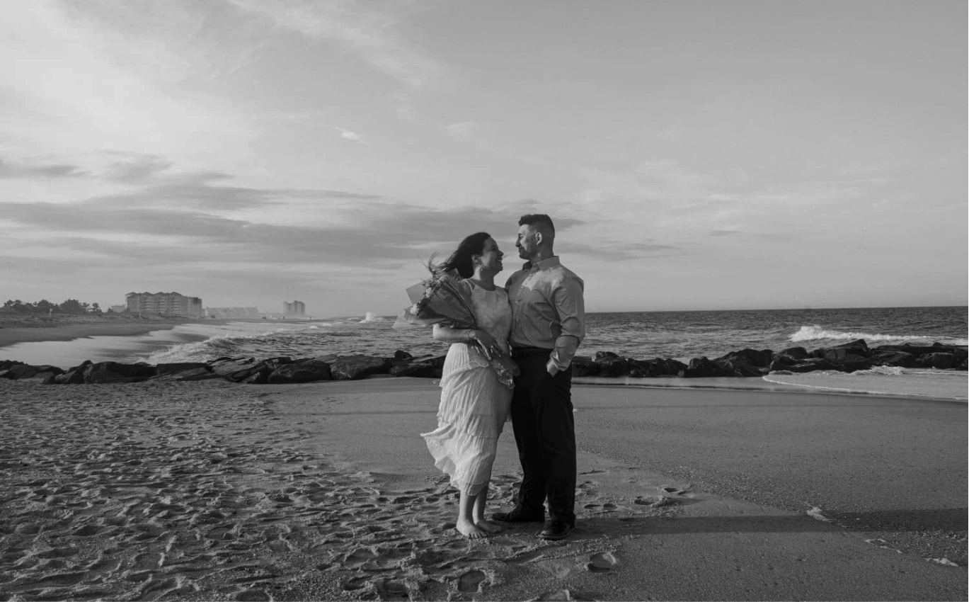 Carlos and Sofia smiling at each other on the beach at sunset, in black and white.